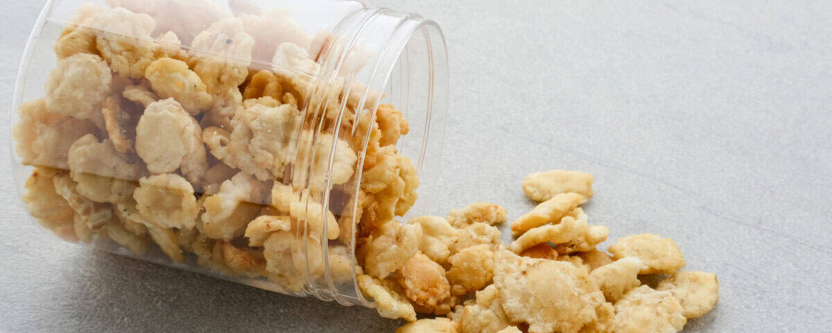 A clear plastic jar tipped over, spilling golden, crunchy Emping Melinjo snacks onto a gray surface. | Emping SG