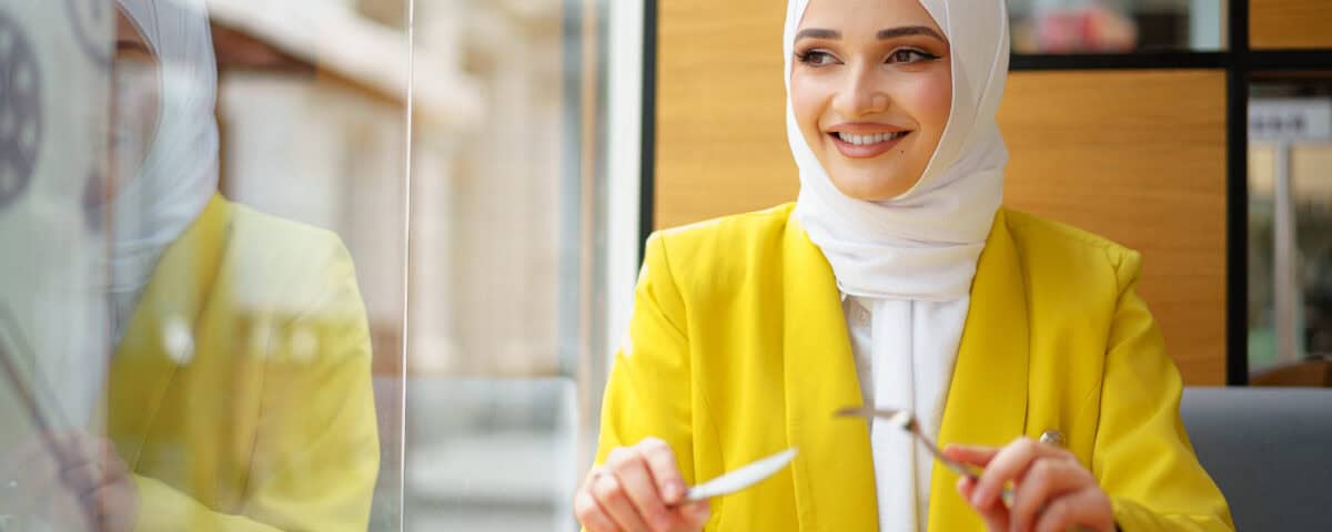 Smiling woman in a yellow blazer and white hijab enjoys a traditional snack at the table, holding a fork and knife. | Emping SG
