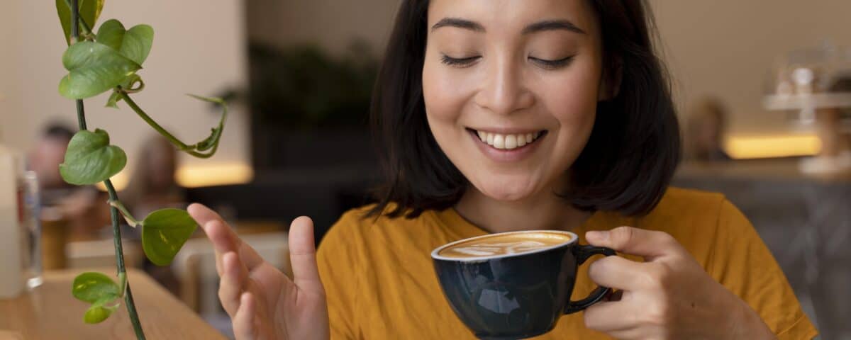 Woman in a yellow shirt smiling and holding a cup of coffee, sitting indoors near a green plant. | Emping SG