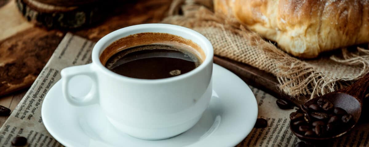 A cup of black coffee on a saucer, with a croissant and coffee beans on a newspaper background. | Emping SG