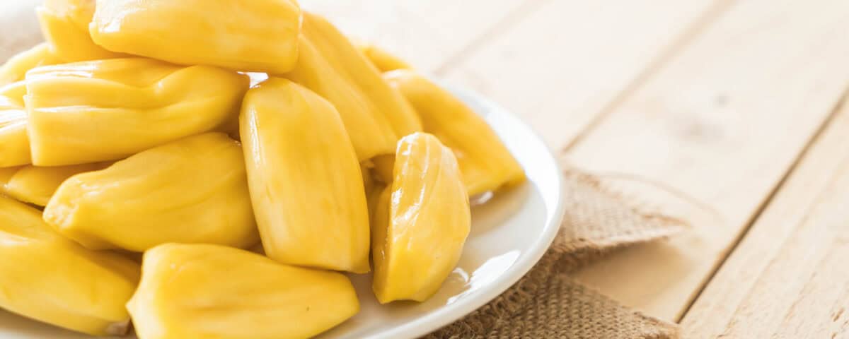 Plate of fresh yellow jackfruit pods, known as Artocarpus heterophyllus, on a white plate atop burlap on a wooden table. | Emping SG