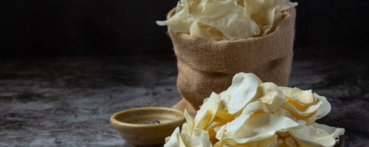 A bowl of thin Emping Melinjo chips, a classic Indonesian snack, sits beside a burlap bag on a bamboo mat. | Emping SG