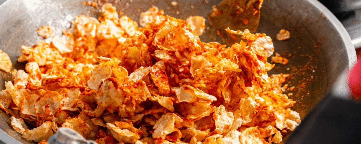 A large metal pan filled with spicy, orange-seasoned potato chips and emping crackers being stirred with a metal spatula. | Emping SG