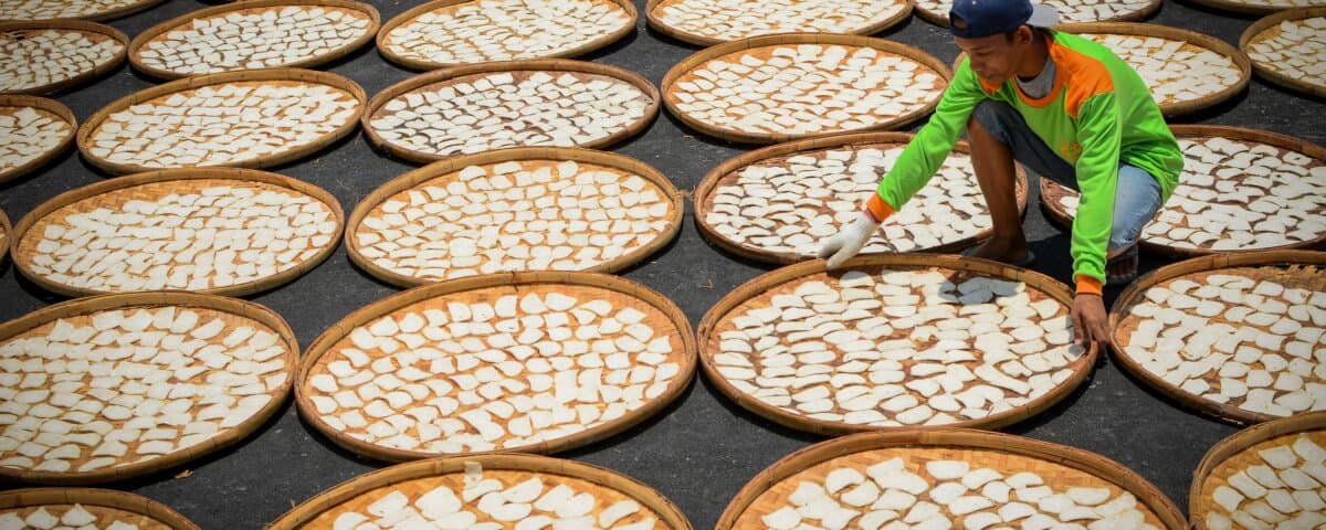 A person arranges round trays of emping melinjo, a traditional gluten-free snack, drying in the sun’s repeating pattern. | Emping SG