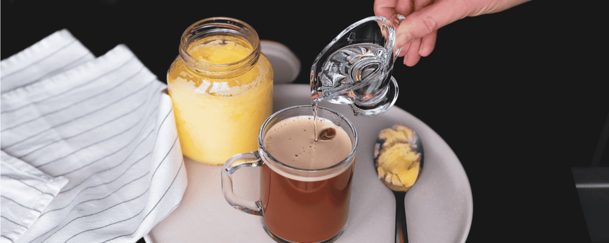 A hand pours liquid into coffee beside ghee, a spoonful of ghee, and a striped cloth—perfecting bulletproof coffee. | Emping SG
