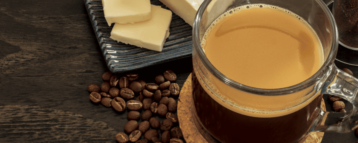 A glass mug of coffee sits next to coffee beans and a small plate with slices of butter on a dark wooden table. | Emping SG