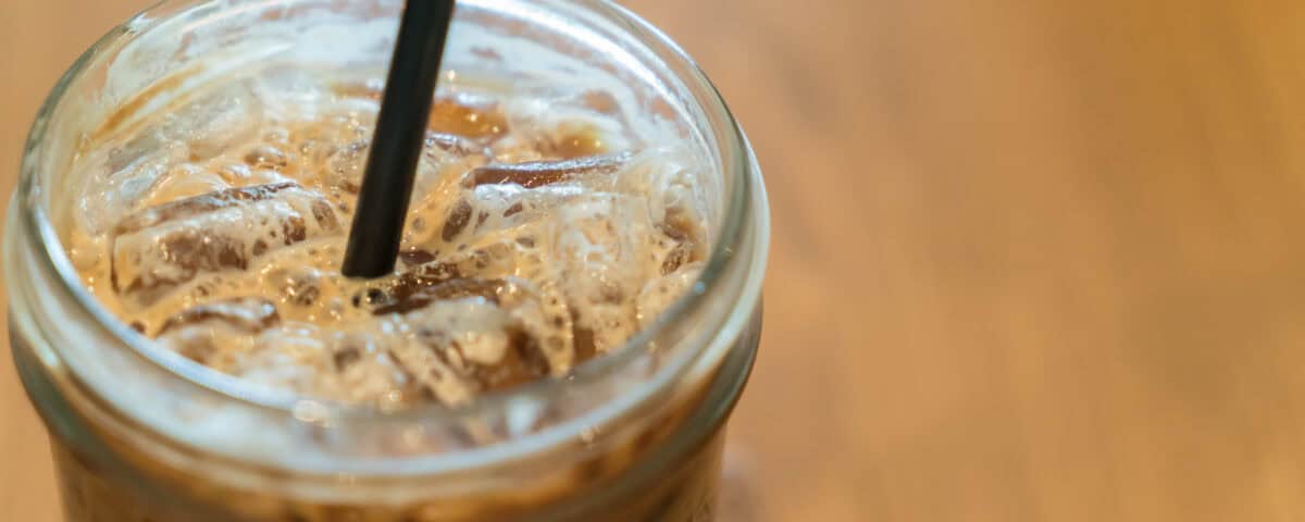 Close-up of an iced coffee in a glass jar with a black straw on a wooden table. | Emping SG