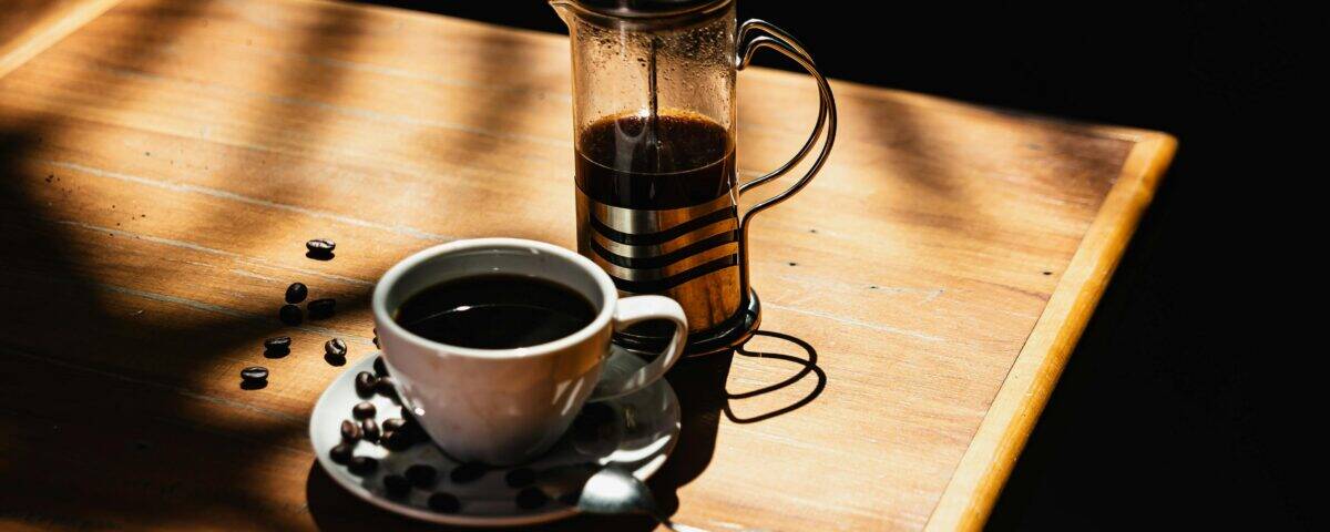 A cup of black coffee with coffee beans, a spoon, and a cafetière on a sunlit wooden table. | Emping SG