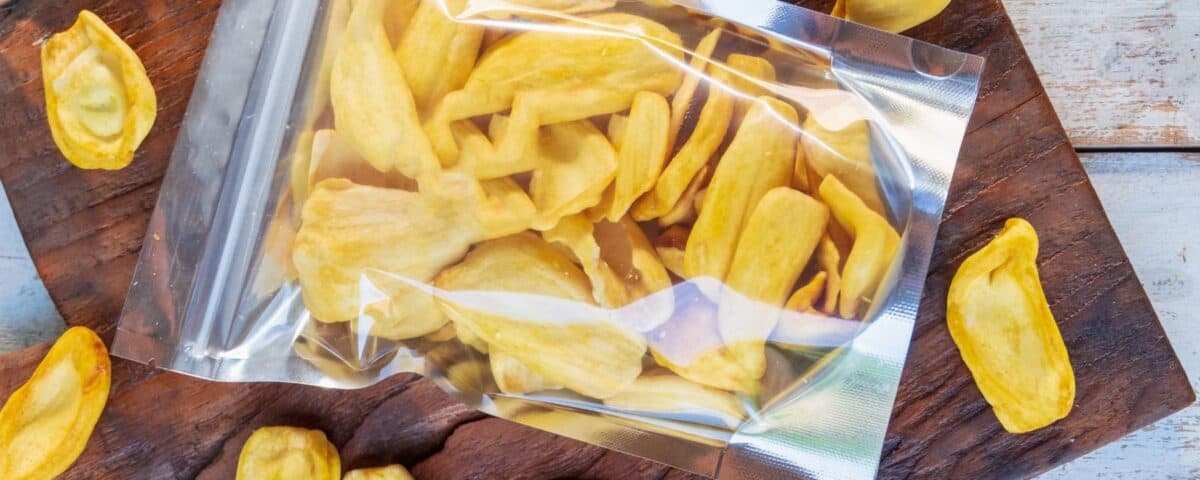 A clear resealable bag of dried jackfruit crisps on a wooden board, with this sweet snack scattered around. | Emping SG