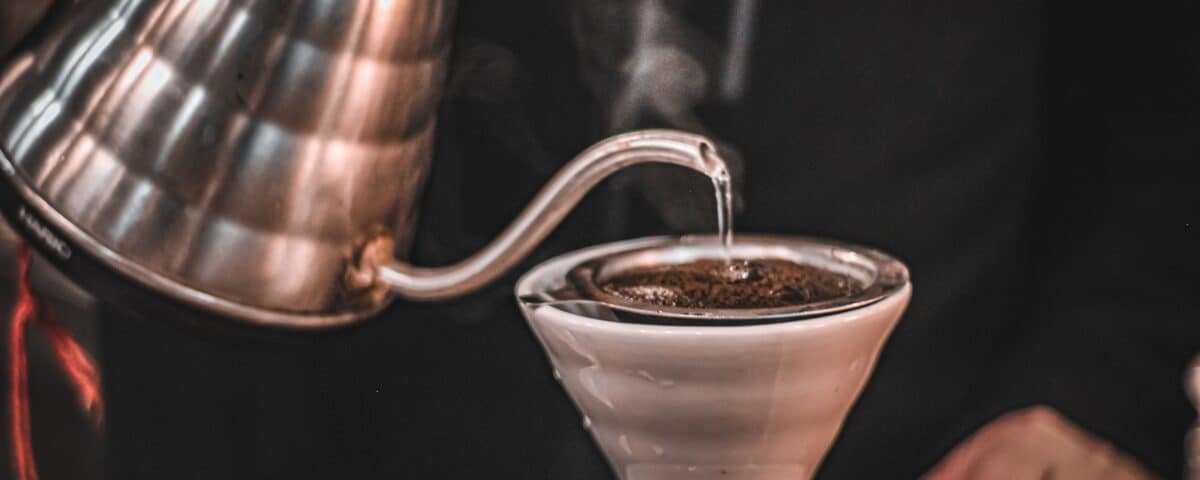 Close-up of hot water being poured from a kettle into a ceramic pour-over coffee dripper with fresh grounds. | Emping SG