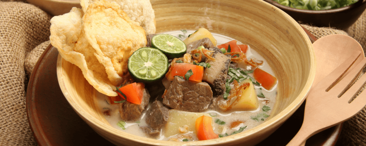 A bowl of Soto Betawi soup with beef, potatoes, tomatoes, lime halves, and crispy crisps. | Emping SG