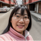 A smiling woman with glasses and long dark hair, wearing a pink jacket, stands on a bustling street with cars and buildings in the background, as if ready to share her testimonial. | Emping SG