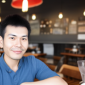 A man in a blue shirt smiles while sitting indoors at a cozy café, with warm lighting and empty tables in the background, seemingly ready to share a glowing review of his delightful experience. | Emping SG