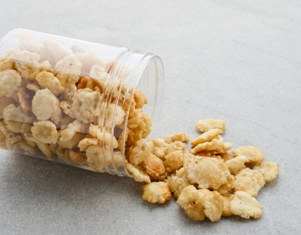 A clear plastic jar tipped over, spilling golden, crunchy Emping Melinjo snacks onto a gray surface. | Emping SG