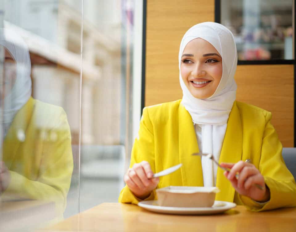 Smiling woman in a yellow blazer and white hijab enjoys a traditional snack at the table, holding a fork and knife. | Emping SG