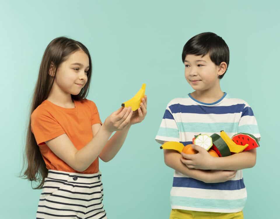 Two kids hold toy fruits, promoting healthy snacks—perfect inspiration for parents and kids. Teal background. | Emping SG