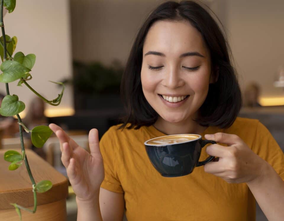 Woman in a yellow shirt smiling and holding a cup of coffee, sitting indoors near a green plant. | Emping SG