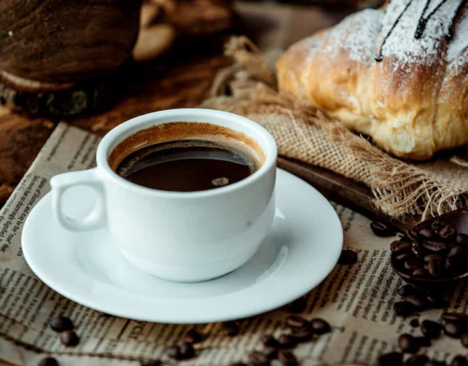 A cup of black coffee on a saucer, with a croissant and coffee beans on a newspaper background. | Emping SG