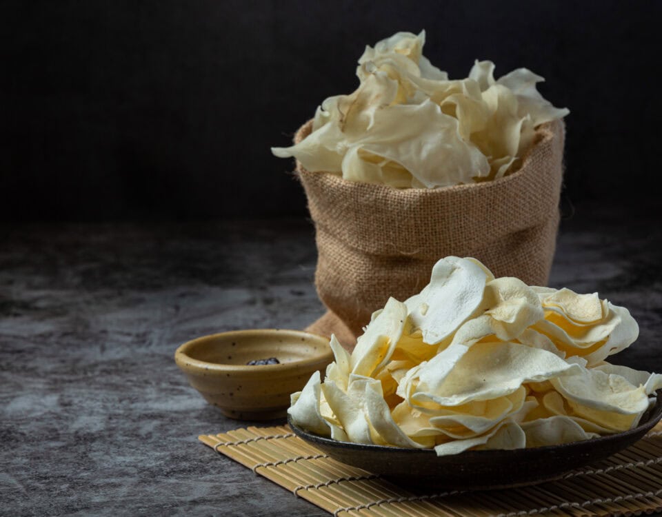 A plate and a burlap sack filled with crispy Emping Belinjo chips on a bamboo mat, with a small dipping bowl nearby. | Emping SG