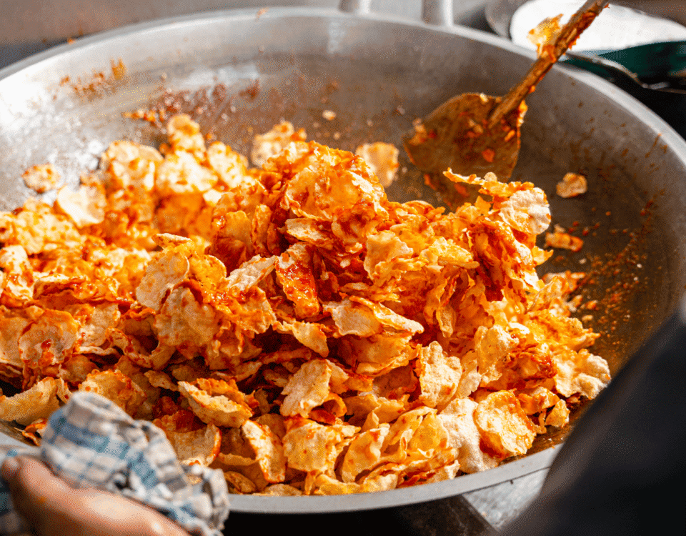 A large metal pan filled with spicy, orange-seasoned potato chips and emping crackers being stirred with a metal spatula. | Emping SG
