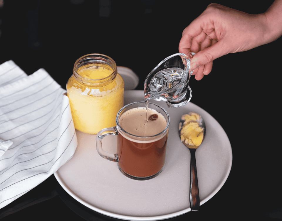 A hand pours liquid into coffee beside ghee, a spoonful of ghee, and a striped cloth—perfecting bulletproof coffee. | Emping SG