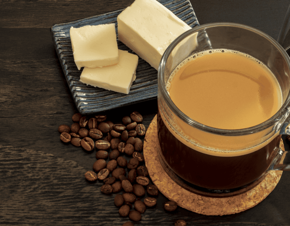 A glass mug of coffee sits next to coffee beans and a small plate with slices of butter on a dark wooden table. | Emping SG