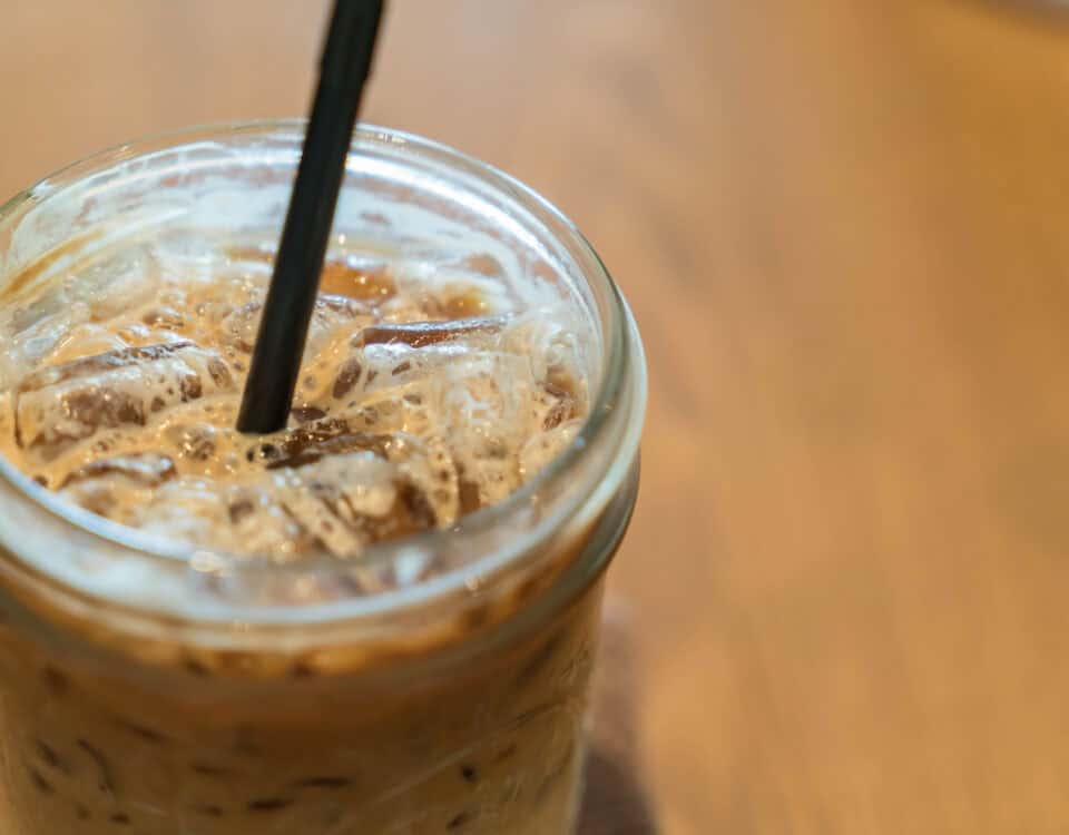 Close-up of an iced coffee in a glass jar with a black straw on a wooden table. | Emping SG