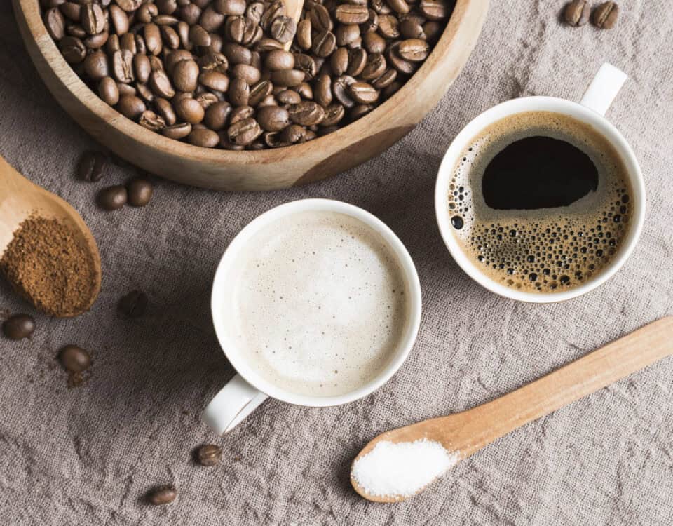 Two cups of coffee, coffee beans, ground coffee, and sugar on a textured cloth with wooden spoons and bowl. | Emping SG