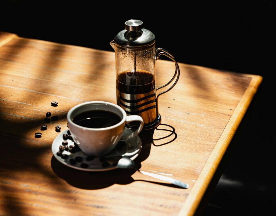 A cup of black coffee with coffee beans, a spoon, and a cafetière on a sunlit wooden table. | Emping SG