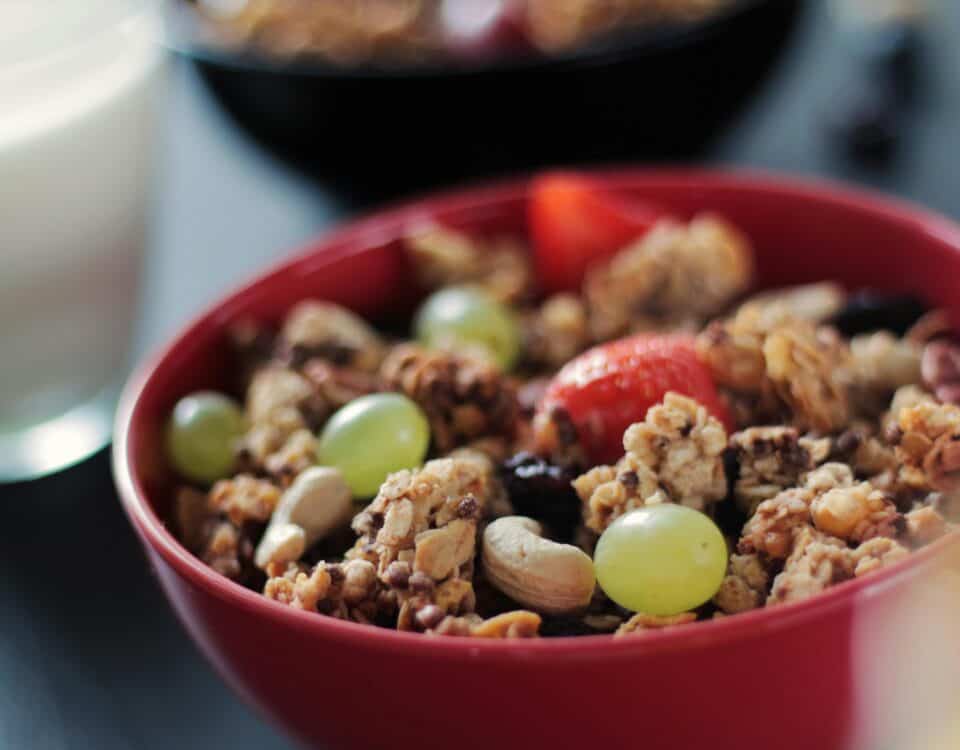 A red bowl filled with high-fibre snacks like granola, grapes, nuts, and strawberries sits next to a glass of milk. | Emping SG