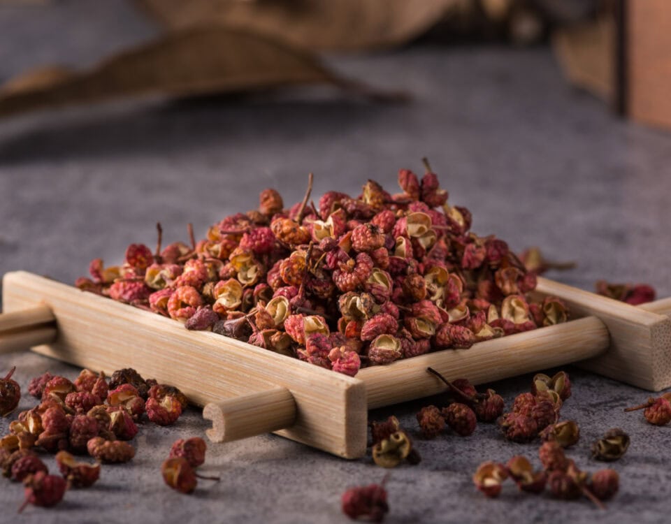 A pile of dried Sichuan peppercorns on a small wooden tray, with some scattered on a grey surface. | Emping SG