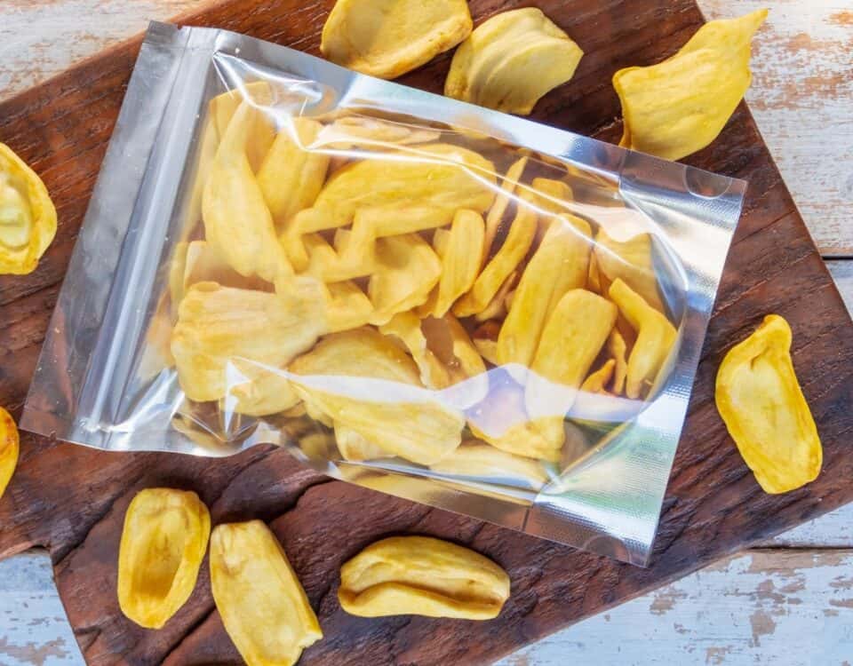 A clear resealable bag of dried jackfruit crisps on a wooden board, with this sweet snack scattered around. | Emping SG