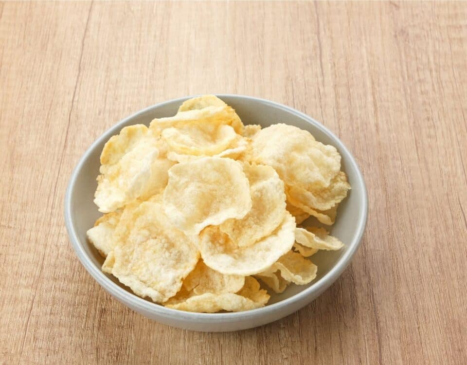 A white bowl filled with crispy emping belinjo crisps on a light wooden surface. | Emping SG