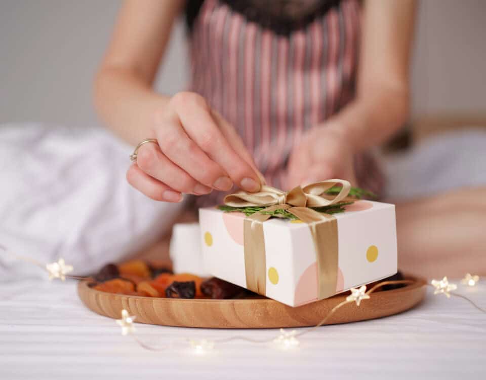 A person unwraps a gift box labelled Healthy Snack Gift Ideas, surrounded by dried fruits and fairy lights on the bed. | Emping SG
