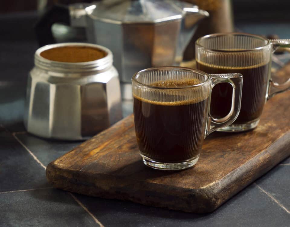 Two glass mugs of fresh roasted coffee on a wooden board beside a moka pot and open coffee tin. | Emping SG