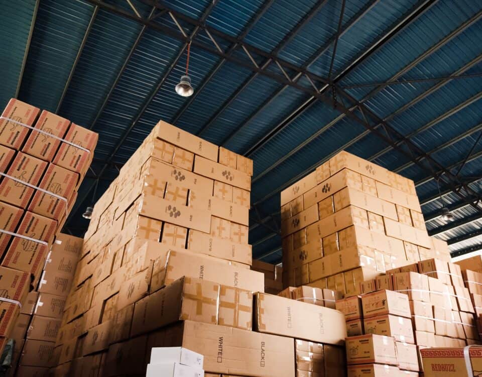 Stacks of cardboard boxes in a warehouse with a metal roof and visible ceiling lights. | Emping SG
